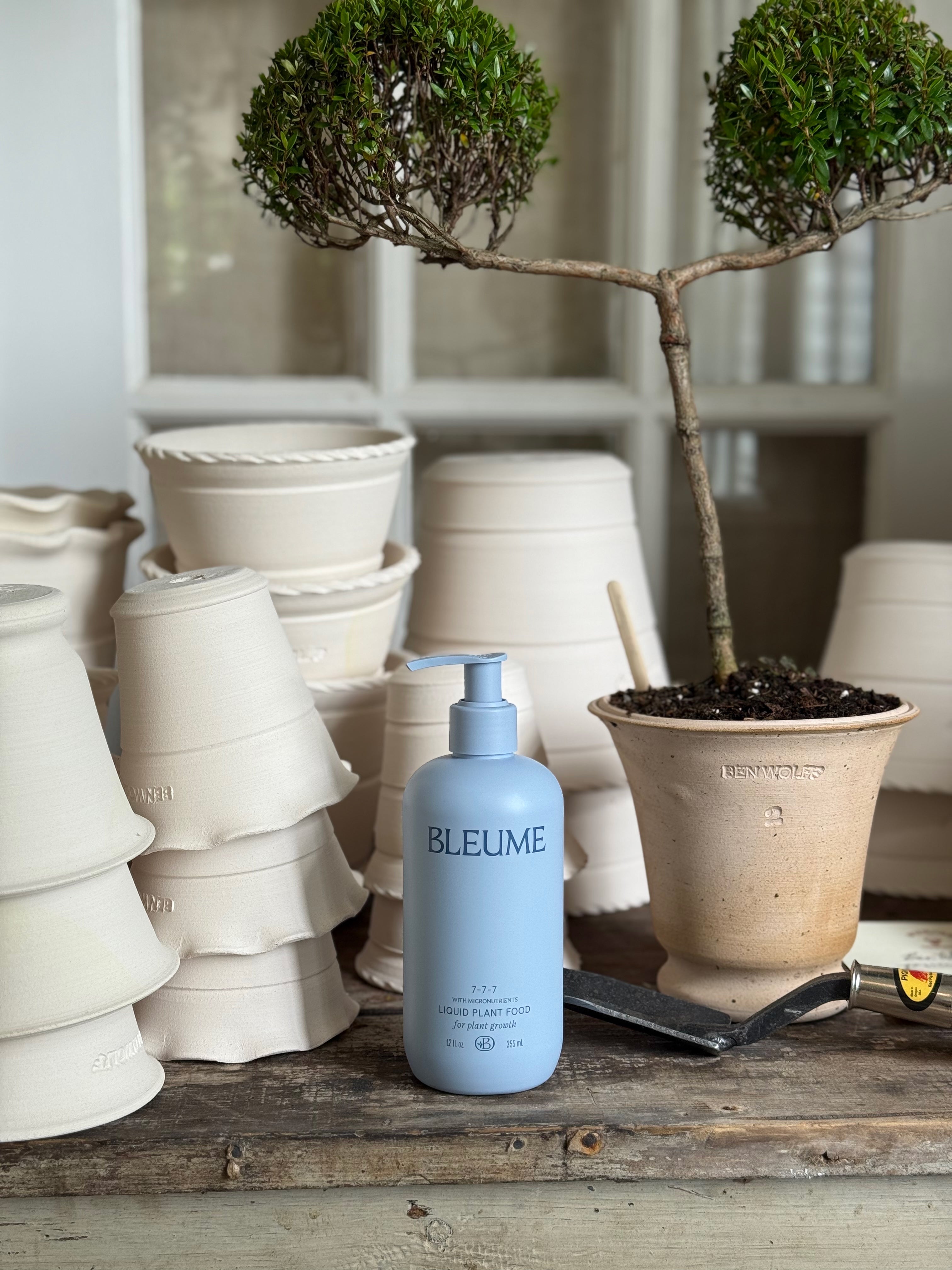 Blue bottle labeled 'BLEUME' on a wooden surface with ceramic pots and plants in the background.
