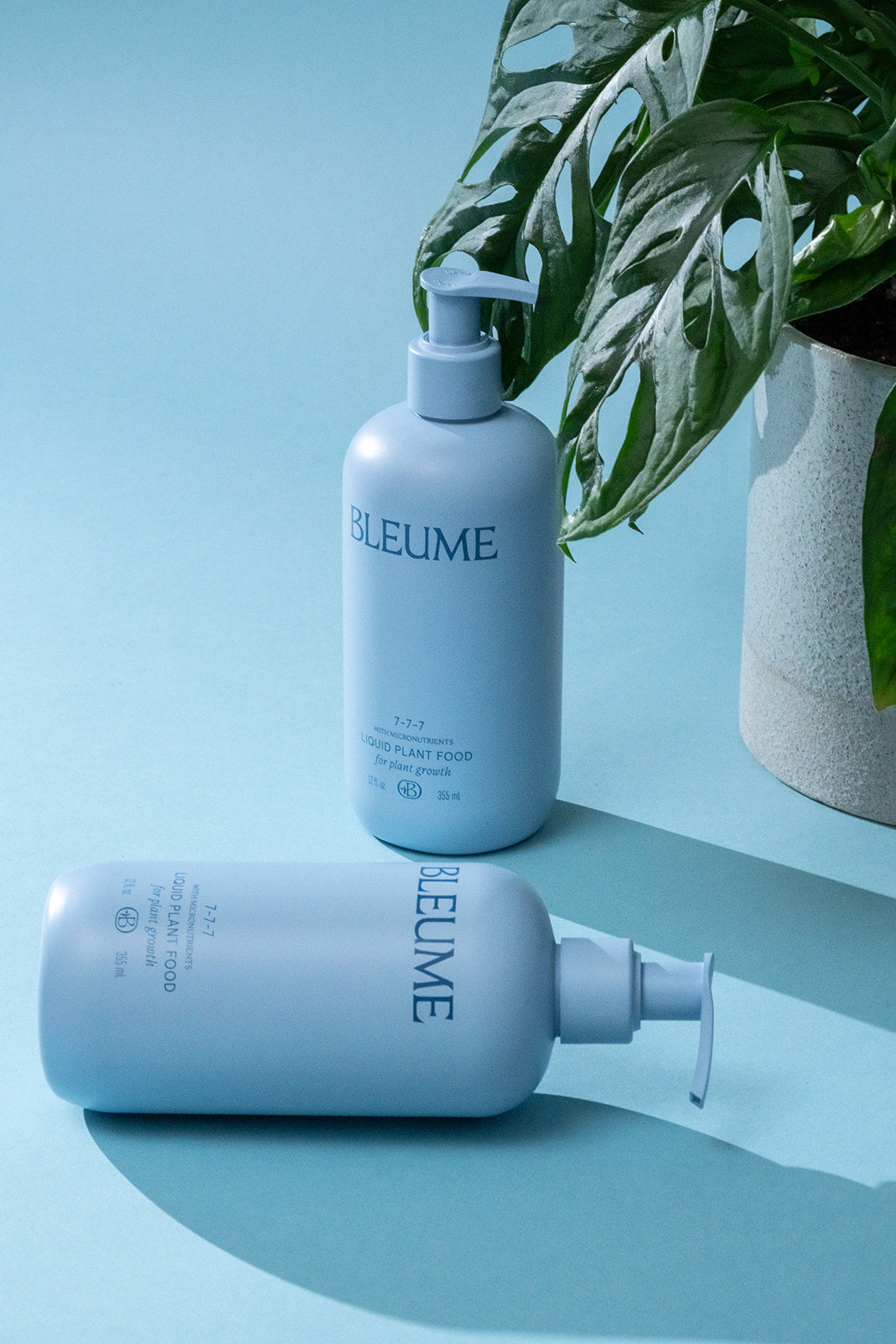 2 blue bottles labeled Bleume on a blue background with a potted plant in the background